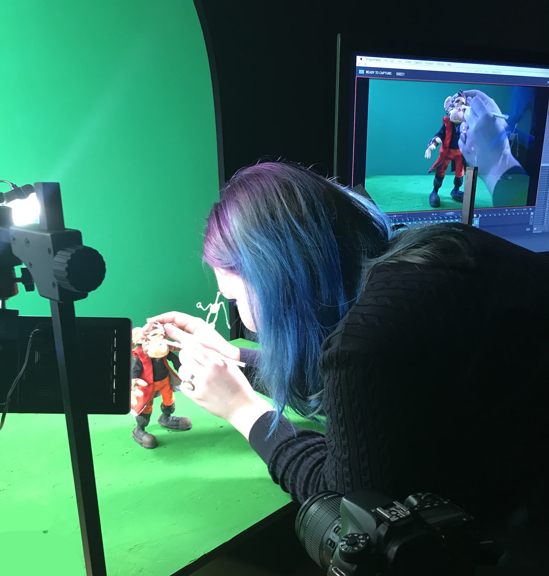 Person with blue hair animating small figures on green screen surface, with camera equipment and monitor displaying the scene.