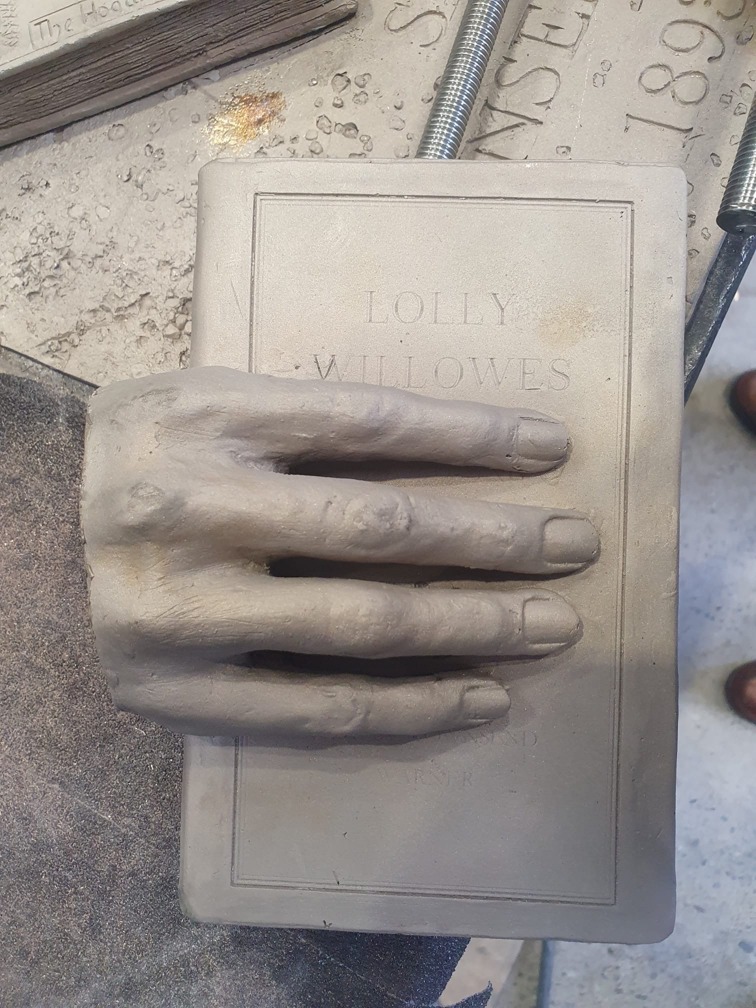 Stone handprint impression in rectangular plaque reading "JOLLY WILLERS" on concrete surface with metal rods visible.