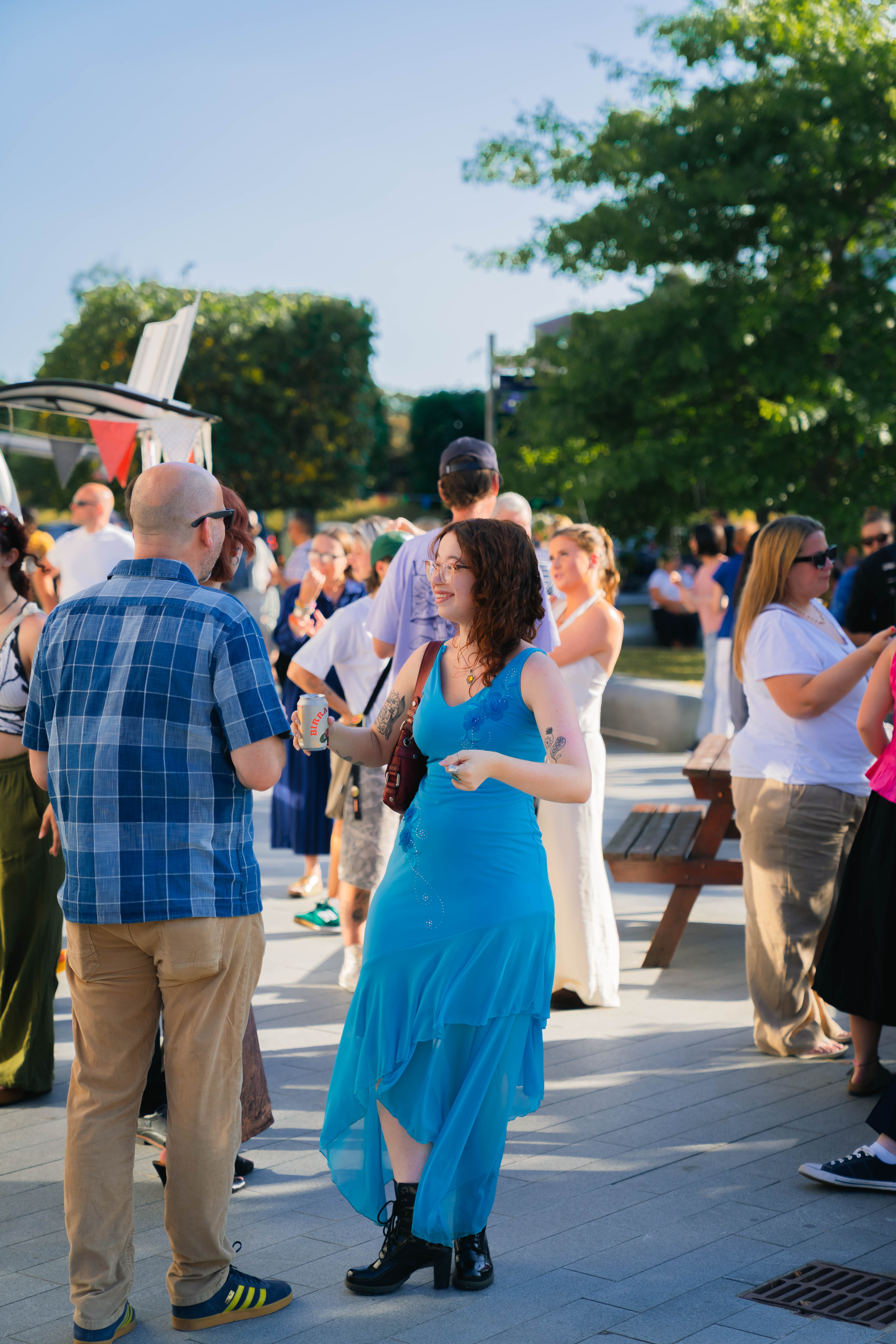 Person stood smiling wearing a vibrant blue dress holding a drinks can talking to someone, people surrounding them talking, sunny evening on AUB Campus with blurred trees in the background.