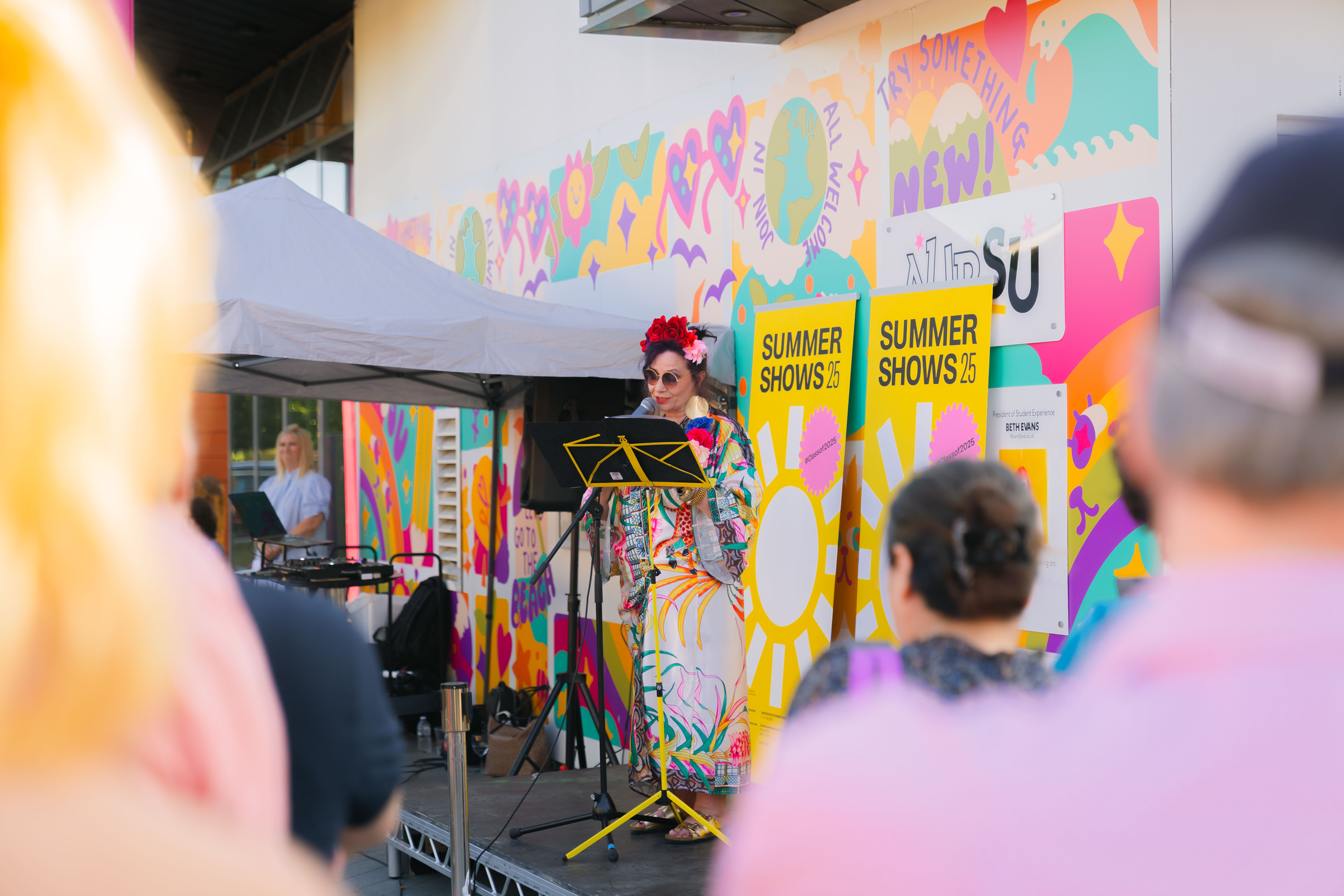 Violet McClean in a multicoloured, floral dress giving a speech in AUB courtyard in front of yellow Summer Show signs and vibrant AUBSU mural.