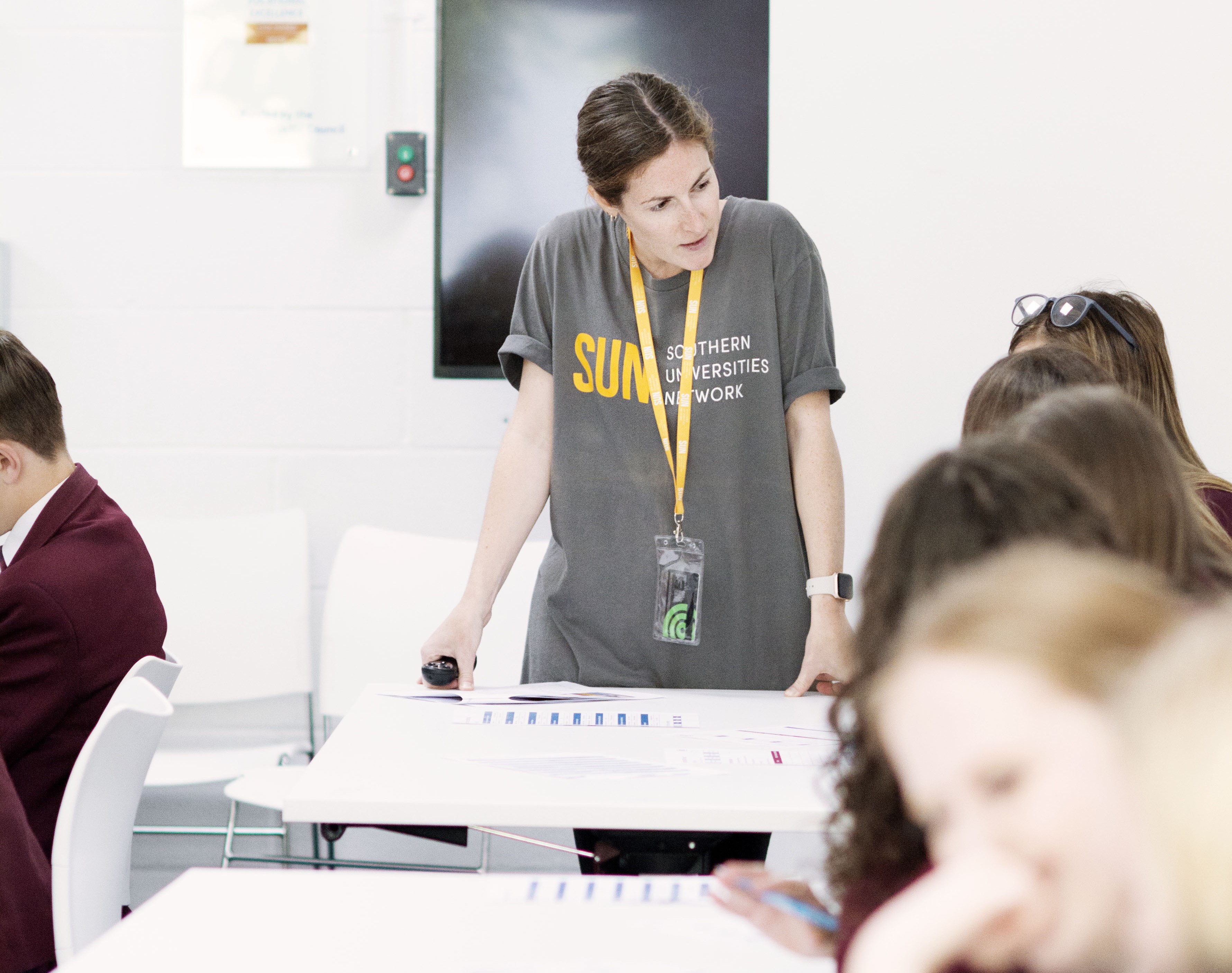 Woman in grey t-shirt with yellow lanyard standing at white table, addressing seated group in classroom setting.