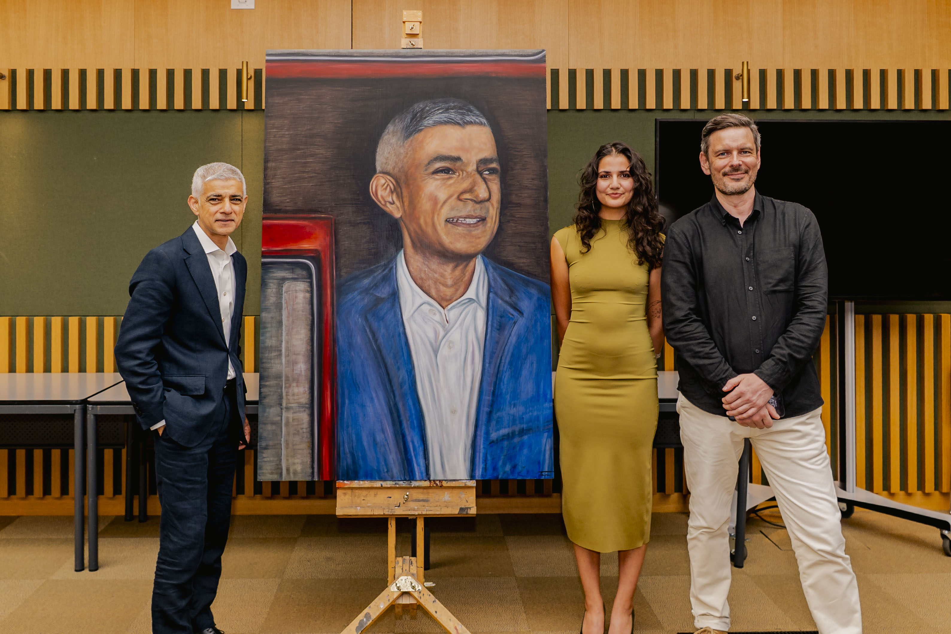 A portrait of Mayor of London Sir Sadiq Khan hangs on an easel, flanked by Sir Sadiq Khan on the left and T Mirza and J Carr on the right. They stand in a room with wooden panelling and large tables.