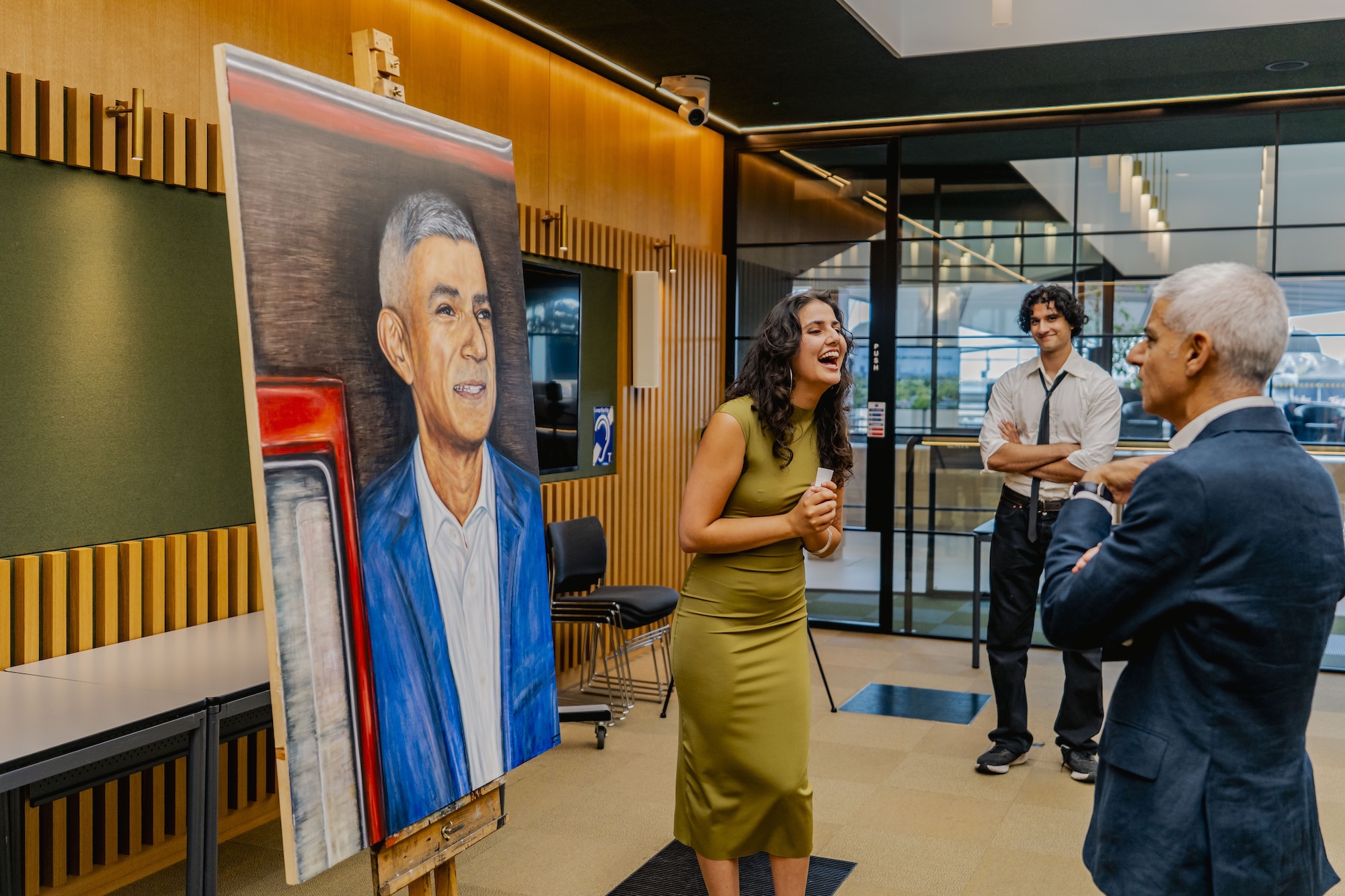 T Mirza stands in front of a large painting of Mayor of London Sir Sadiq Khan. Sir Sadiq Khan is observing the painting with another man standing behind, all in a modern indoor setting with wooden and glass features.
