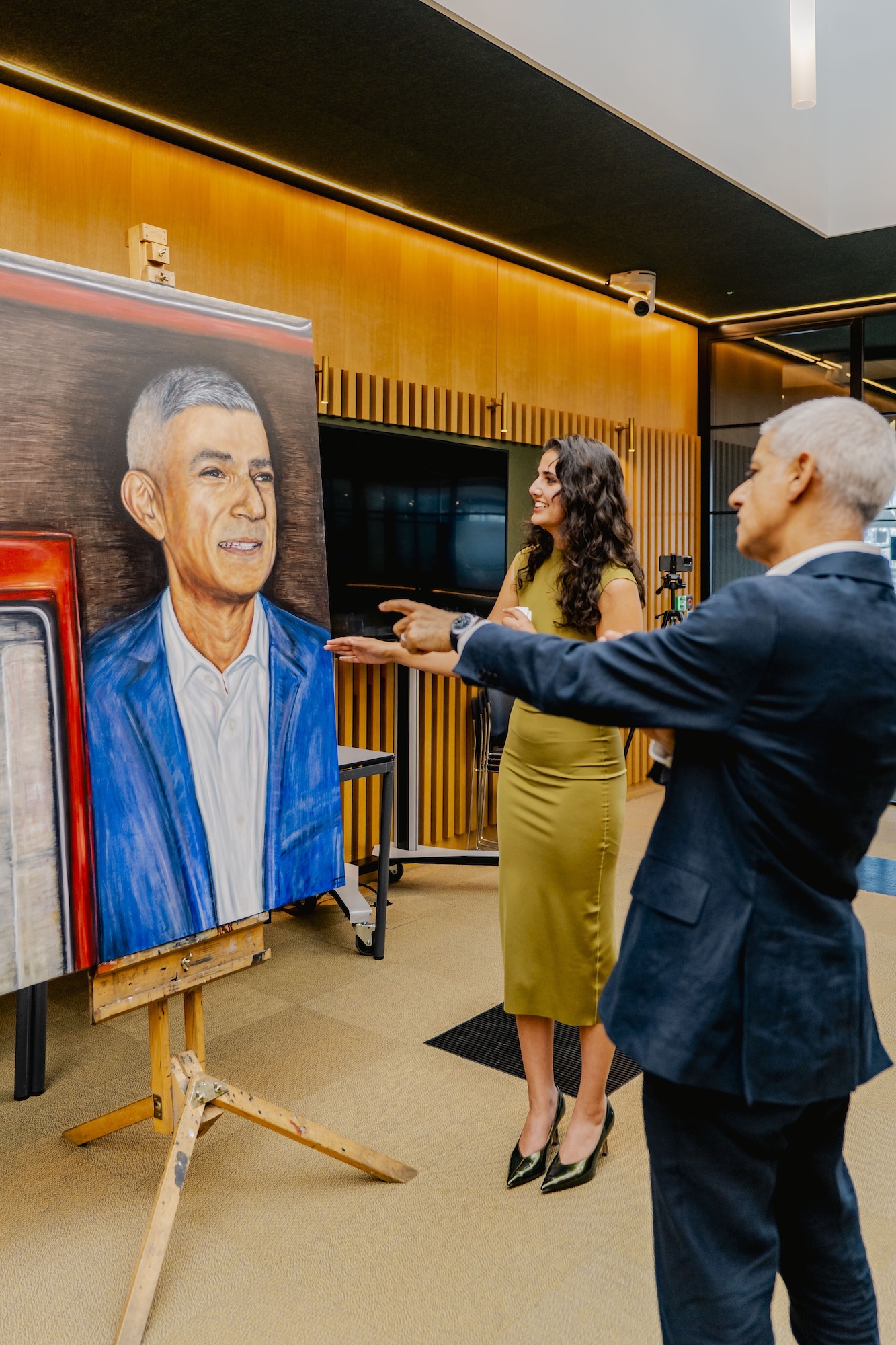 T Mirza stands beside Mayor of London Sir Sadiq Khan, pointing at a portrait on an easel. The portrait displays a man dressed in a blue suit with a white shirt. The background features wooden paneling and modern decor.