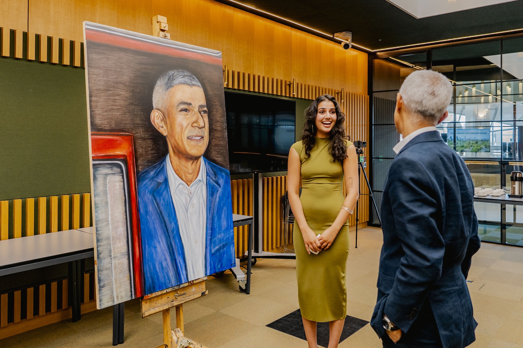 T Mirza stands next to a portrait of Mayor of London Sir Sadiq Khan, displayed on an easel. Sir Sadiq Khan stands with his back to the camera in the foreground.