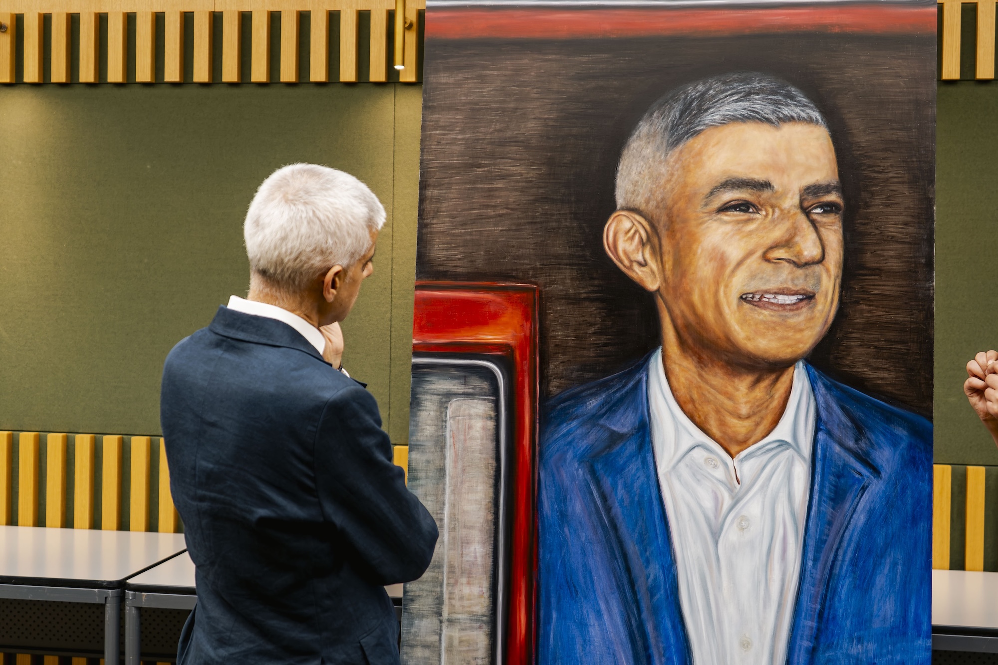 Mayor of London Sir Sadiq Khan with his back to the viewer, observing a large portrait of a man with short grey hair and a blue suit. The background features a mix of green and wooden elements, enhancing the gallery setting.