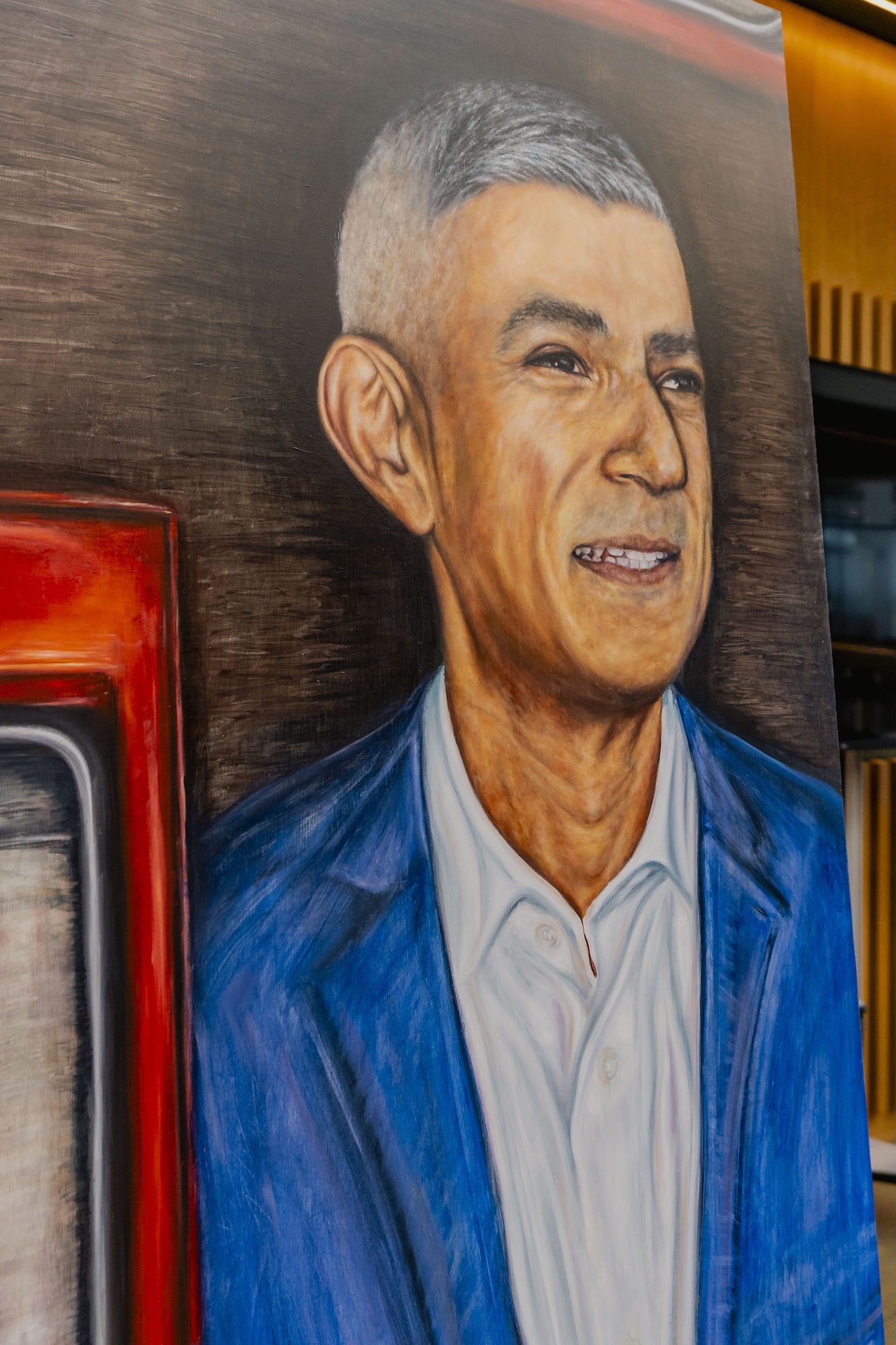 A portrait of Mayor of London Sir Sadiq Khan. He is positioned next to a red element, with a textured brown background behind him.