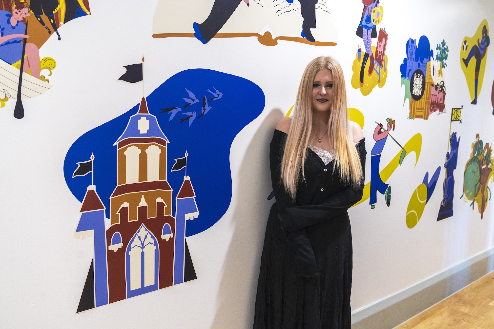 BA (Hons) Illustration student S Newman poses for a photo next to a colourful wall vinyl illustration of a manor or castle-looking building next to them.