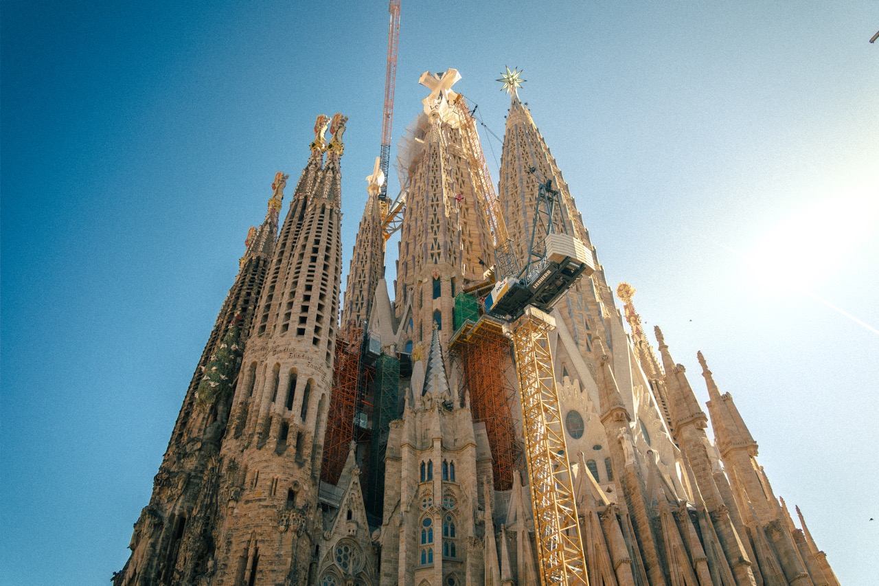 The Sagrada Familia, mutiple, detailed tall towers with cranes and scaffolding, blue sky in the background.