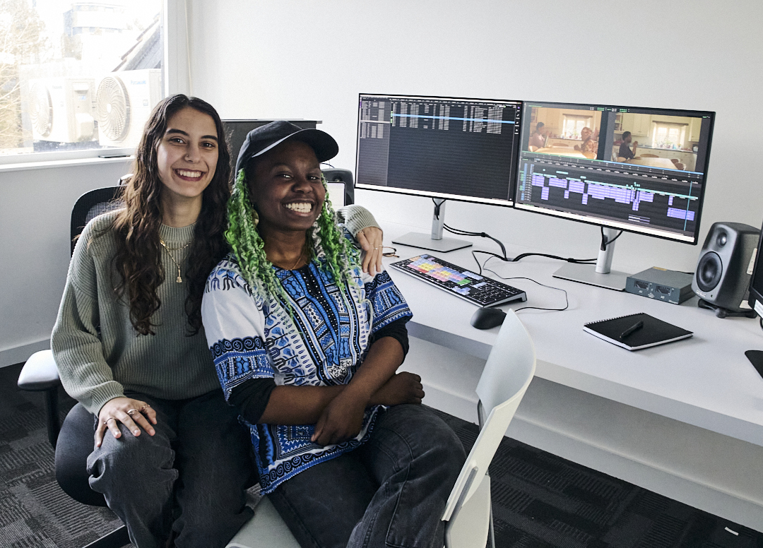 Two women sitting at white desk with dual monitors displaying video editing software, speakers, and tablet in bright office space.