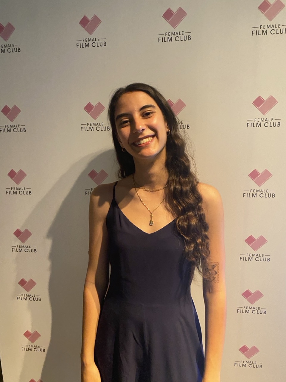 Woman in dark blue dress smiling at camera against white backdrop with pink heart-shaped logos reading "Female Film Club".