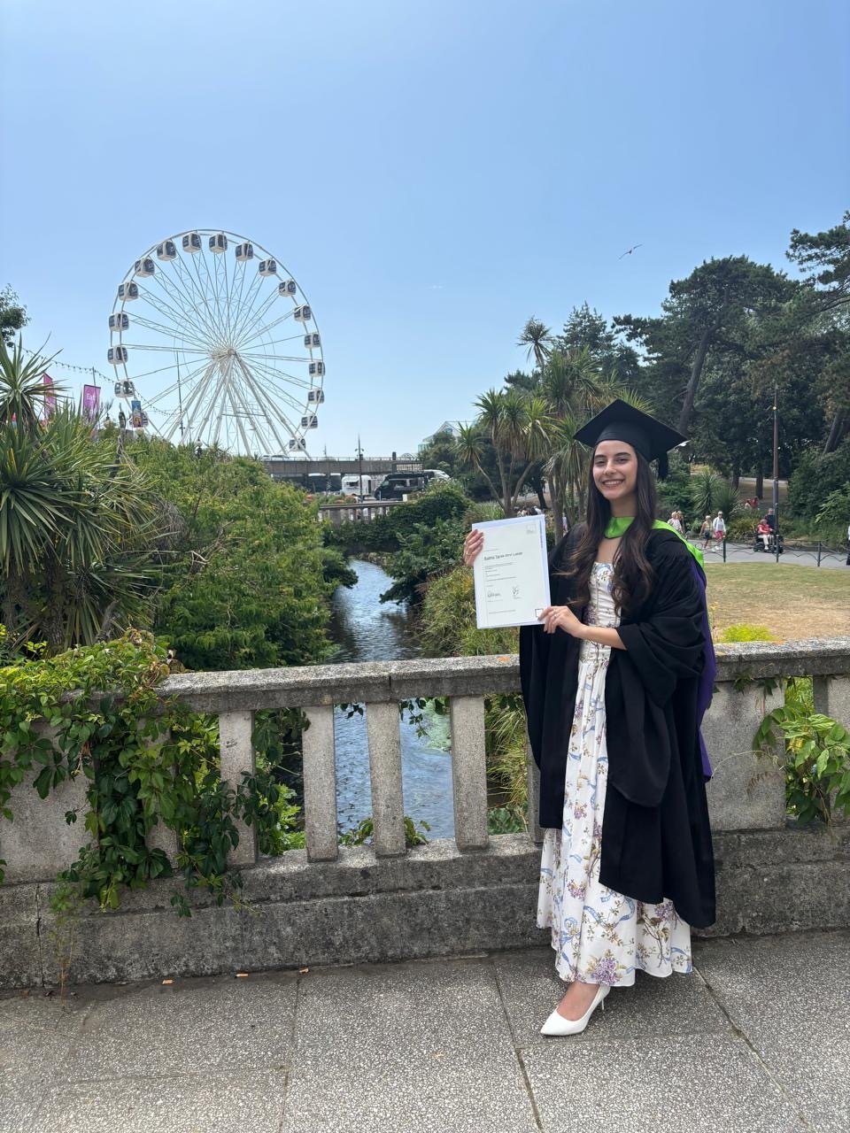 Graduate in black gown and mortarboard holding certificate, standing by stone balustrade with ferris wheel and green parkland behind.