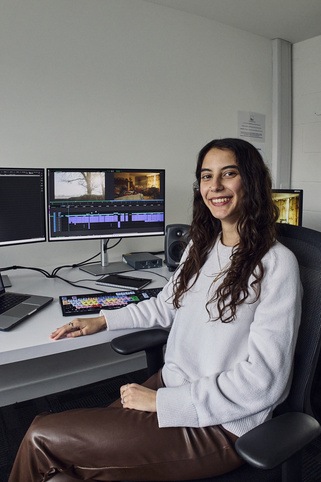 Woman with long dark hair in white jumper smiling at desk with multiple computer monitors showing video editing software.