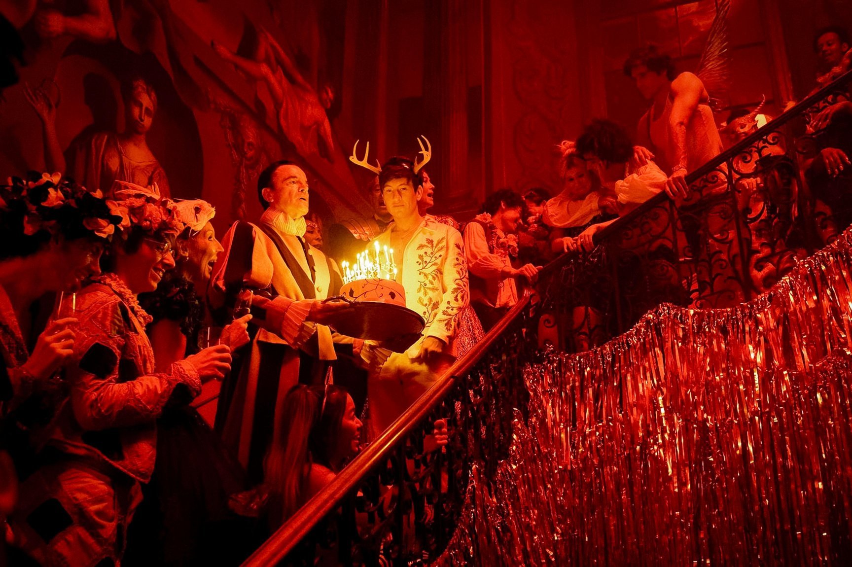 A scene from Saltburn, with character Oliver Quick wearing deer antlers with a birthday cake, surrounded by people on the stairs in red light.