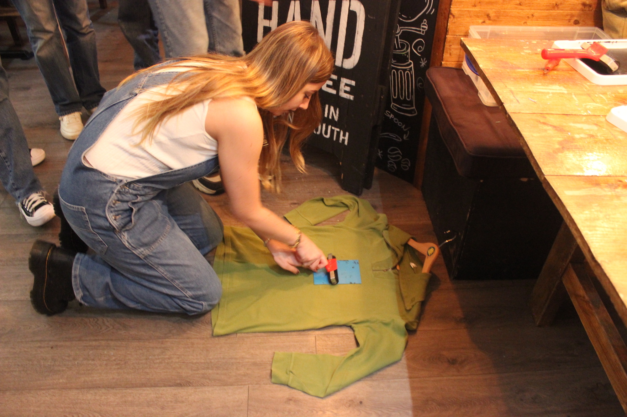A girl kneeling down and using a tool to patch over a garment that is laid out on the floor.