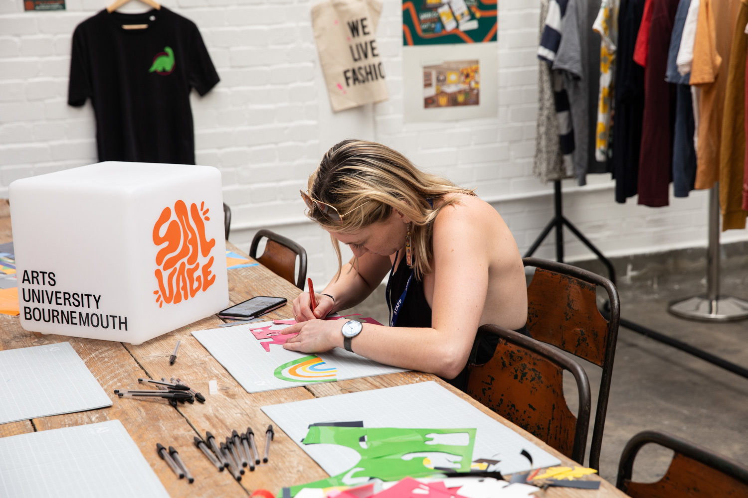 A person sitting at a table working with stencils. A cube sits on the table next to them with the Salvage Collective and Arts University Bournemouth logos on it.