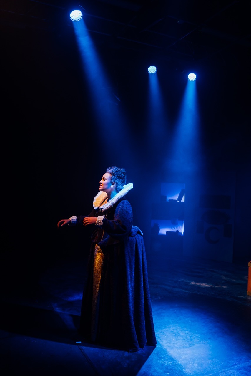 Performer in period costume on stage under three bright blue spotlights against dark background.