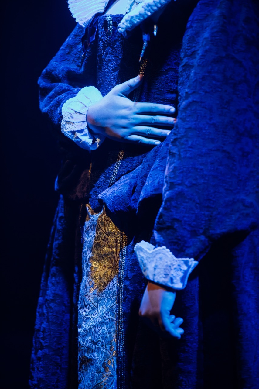 Two hands clasped together in blue lighting, wearing ornate period costume with gold embroidered waistcoat and lace cuffs.
