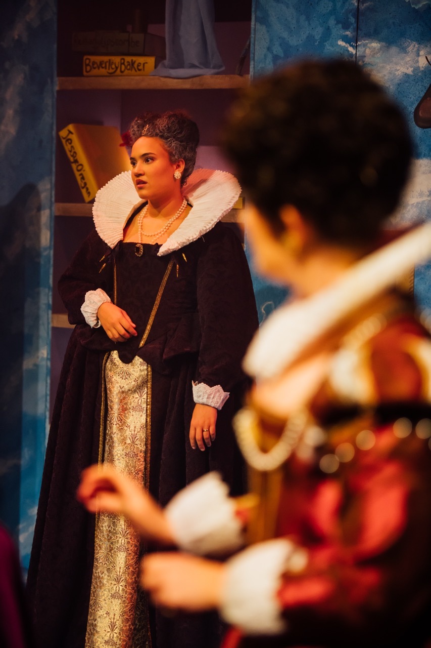 Actor in Elizabethan costume with white ruff collar and crown on stage, with blurred figure in foreground during theatre performance.