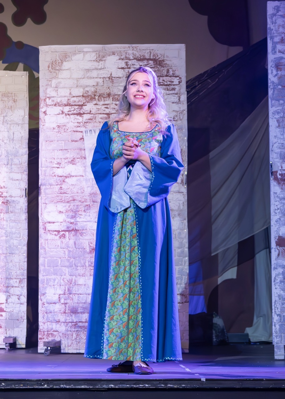 Woman in blue medieval dress with floral bodice standing on stage, stone wall backdrop, theatrical lighting.