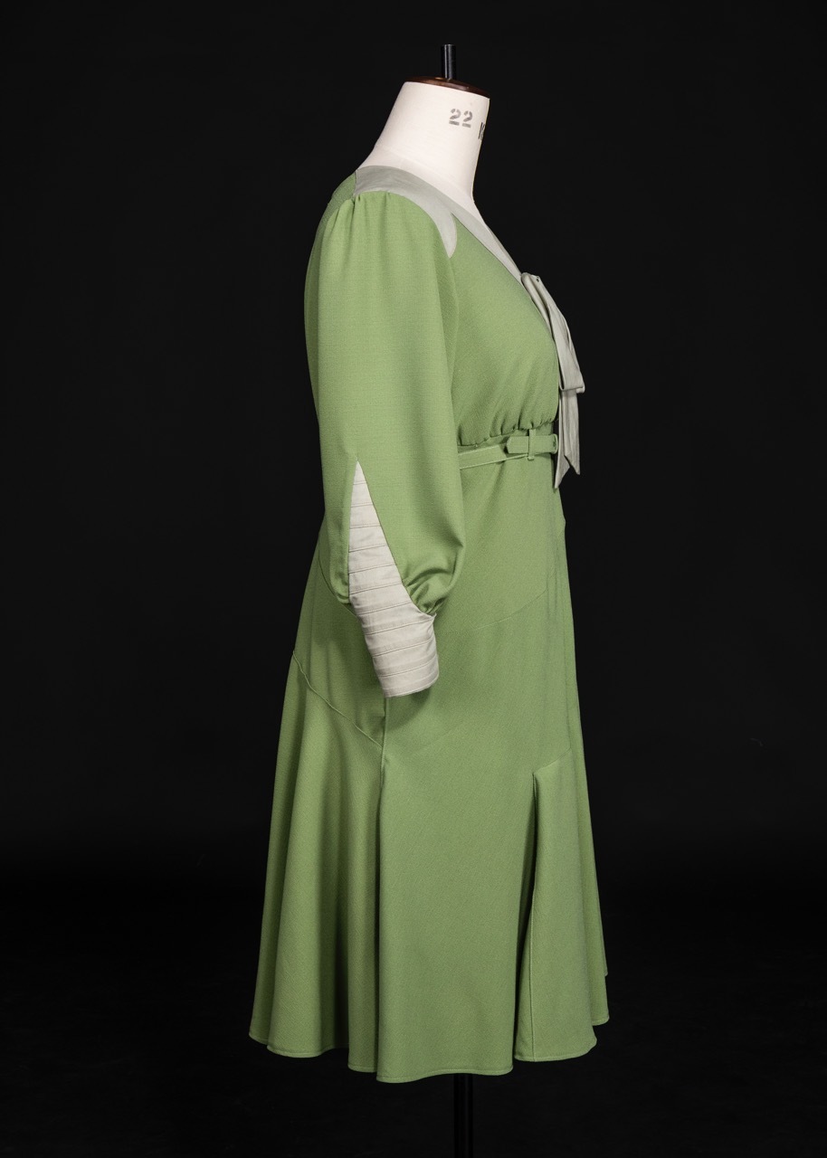 Green dress with long sleeves and white collar displayed on dressmaker's mannequin against black background, shown from side view.