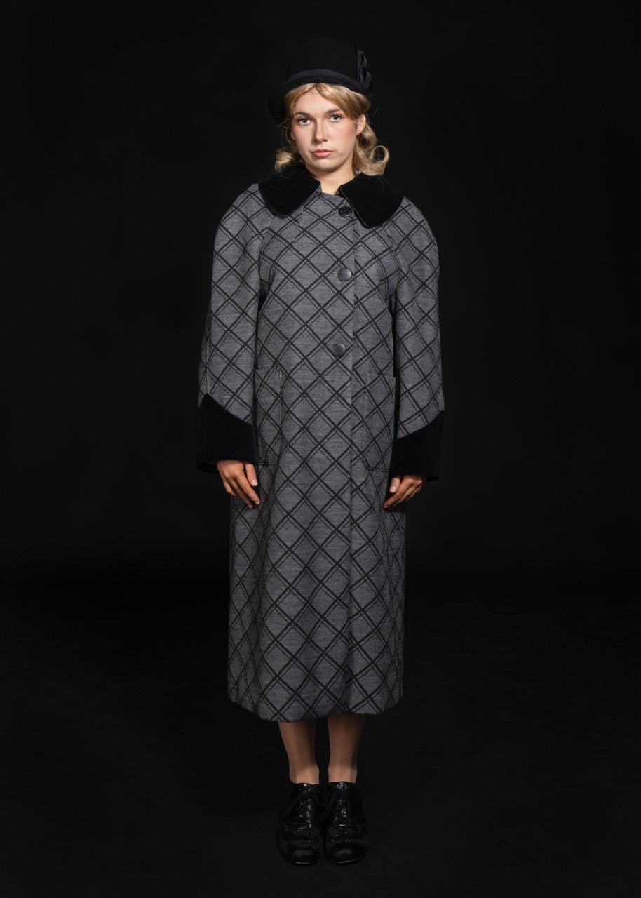 Model on runway wearing grey diamond-patterned midi coat with black collar and cuffs, black boots, blonde hair, dark background.