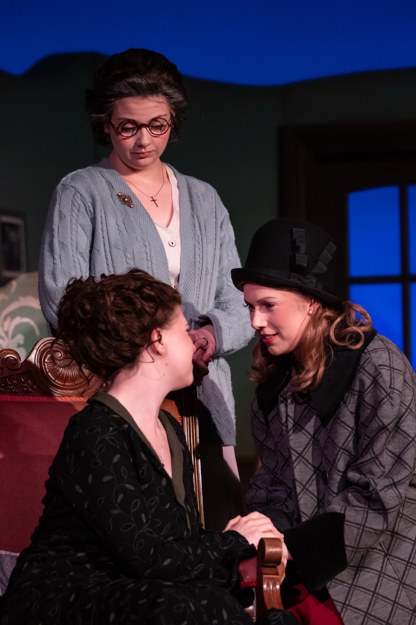 Three women on theatre stage. Woman in grey cardigan and glasses stands behind two seated women - one in black dress, one in grey coat and hat.