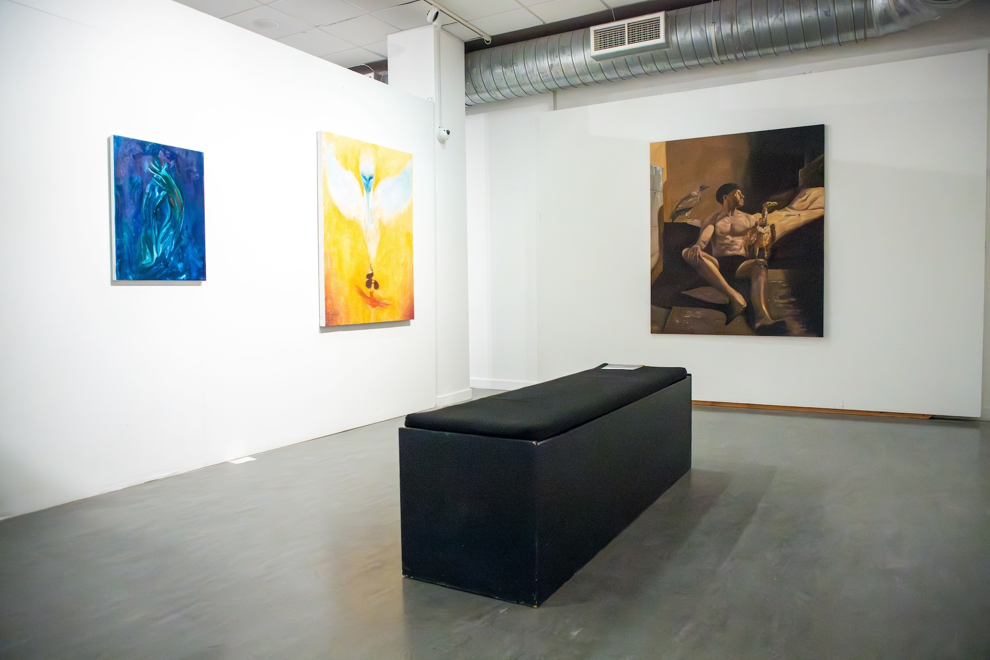 Three paintings, one showing a shirtless male sitting in front of a bed, one of a white wading bird with its wings spread on a golden background, and a primarily blue abstract painting, all on display in a gallery space with a long black seat in the centre.