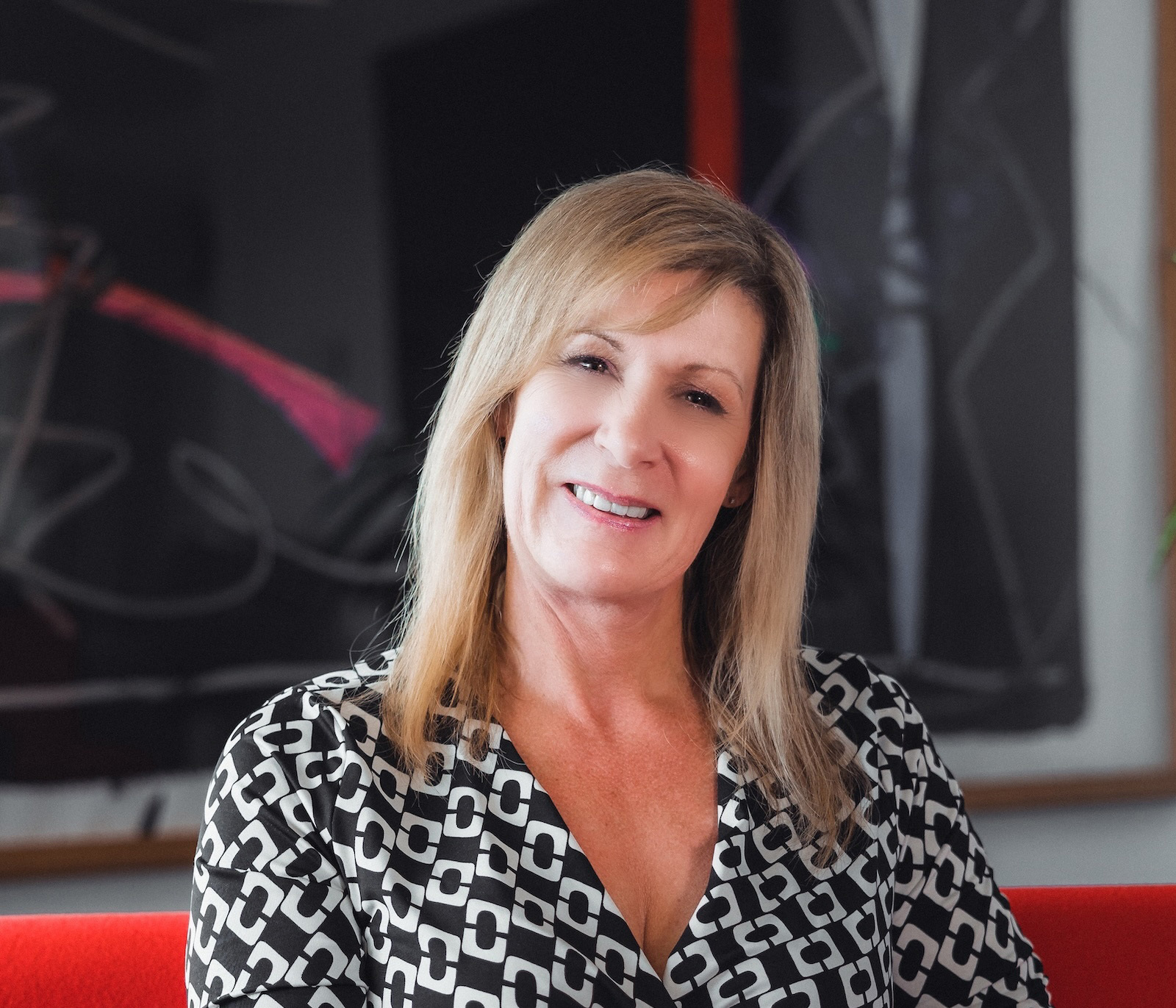 Blonde woman in black and white geometric patterned top smiling at camera, with blurred artwork visible in background.