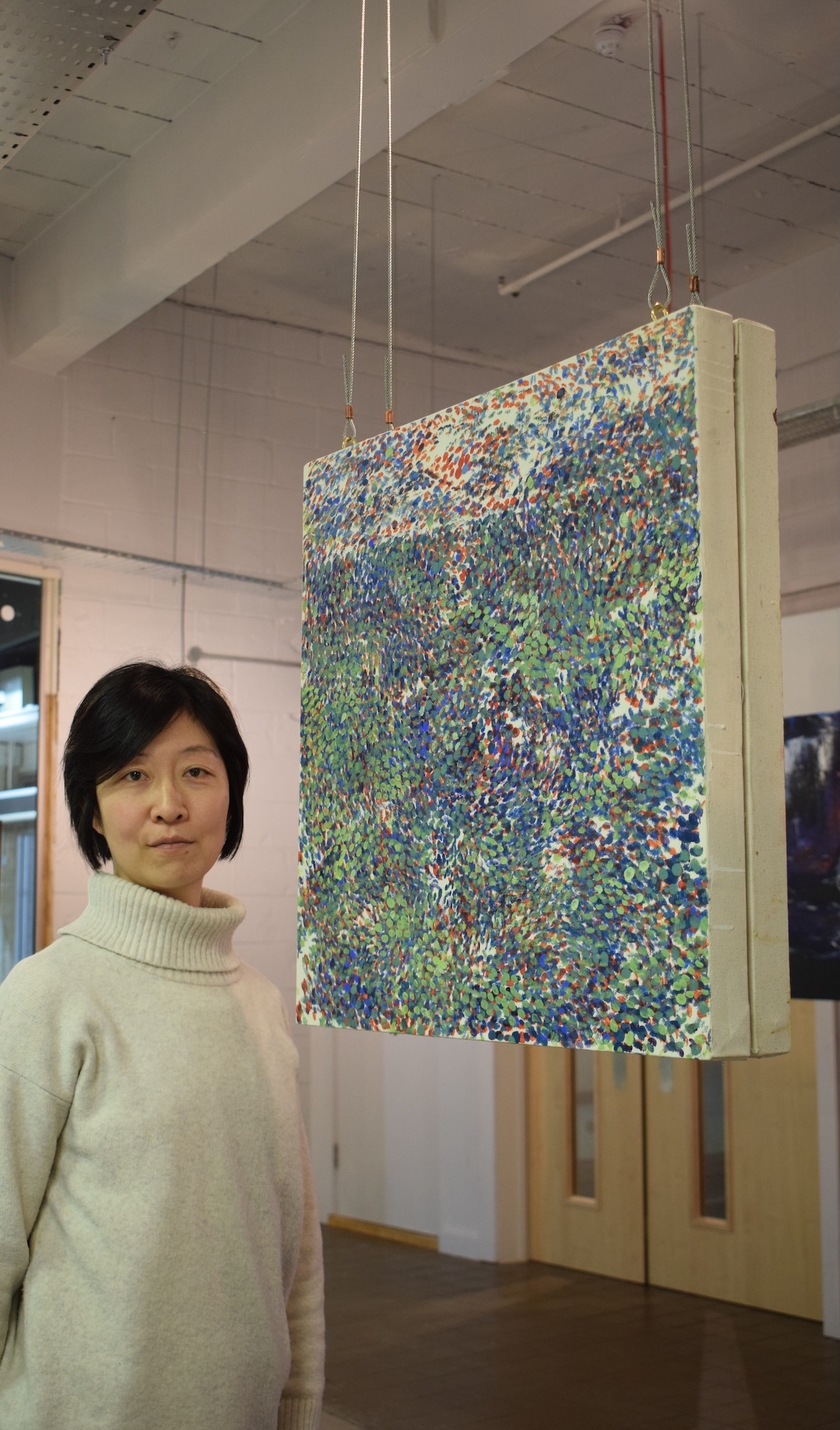S Takahata standing beside suspended artwork with dense multi-coloured abstract pattern in gallery space