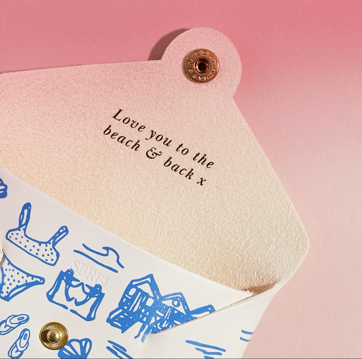 A white pouch with blue illustrations and embroidered text: 'Love you to the beach & back x', pink background.