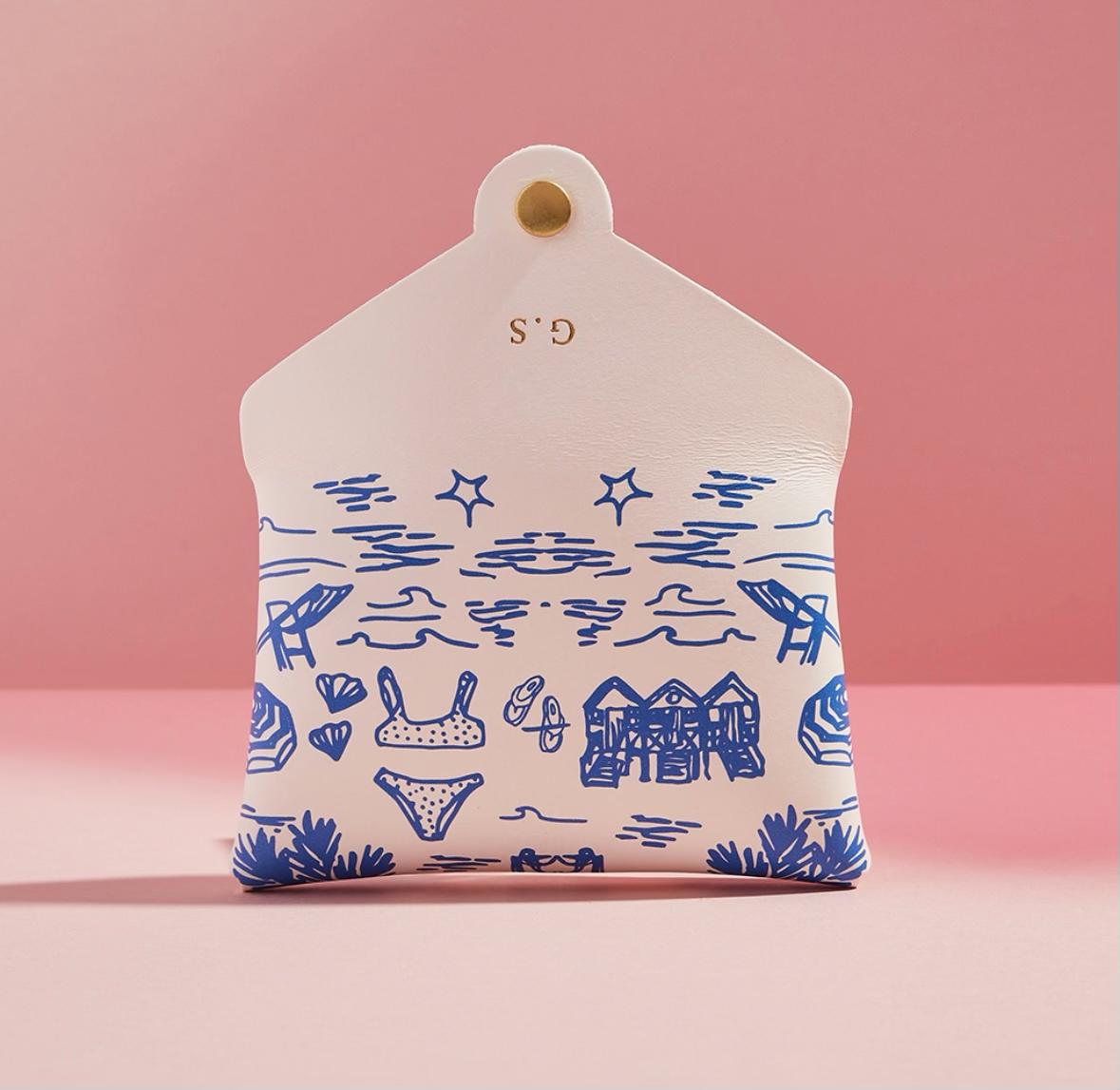 A white pouch with blue illustrations and gold embroidered initials 'G.S.', pink background.