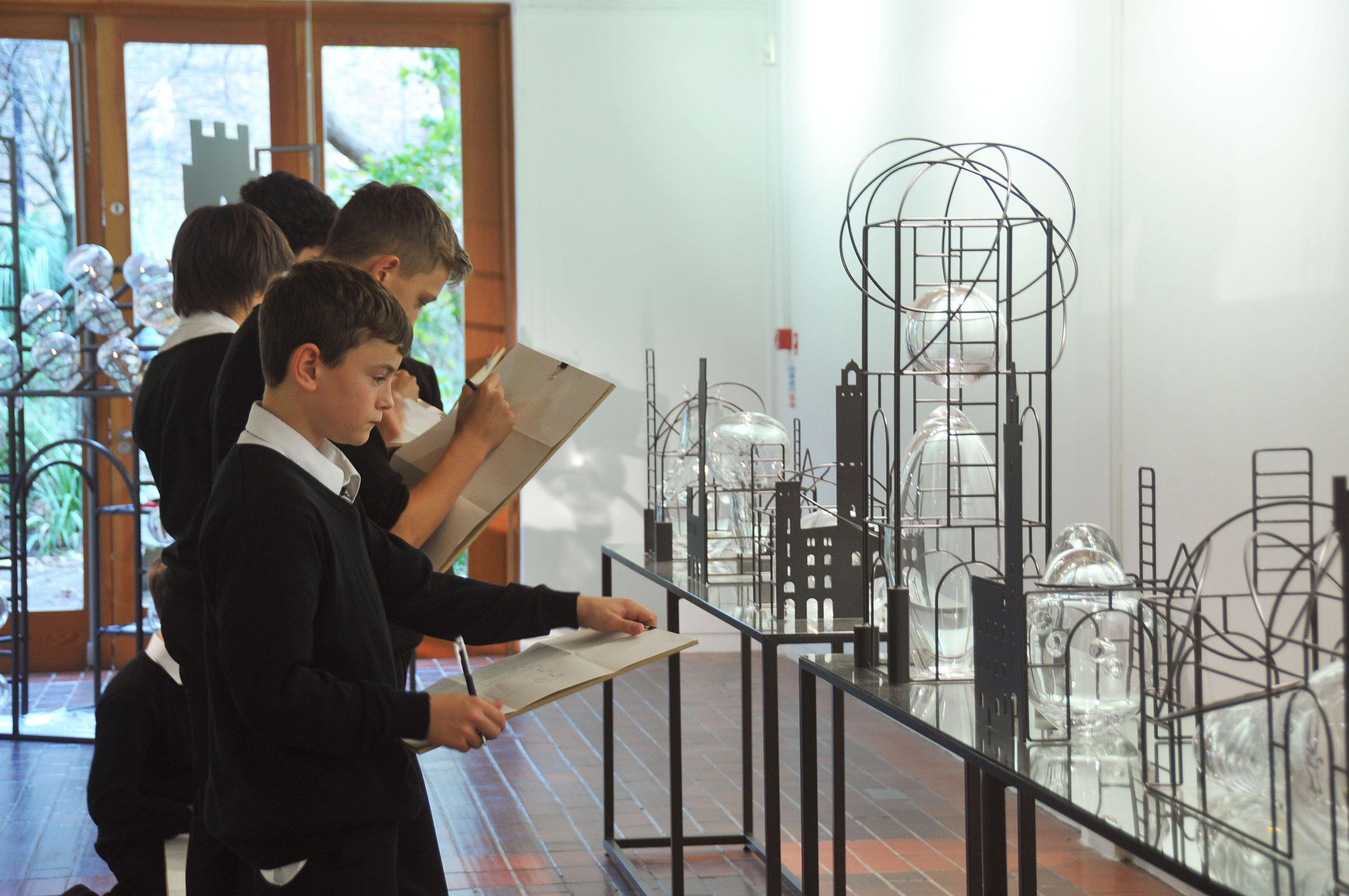 A line of children holding clipboards and observing a set of sculptures on display on tables in a gallery.