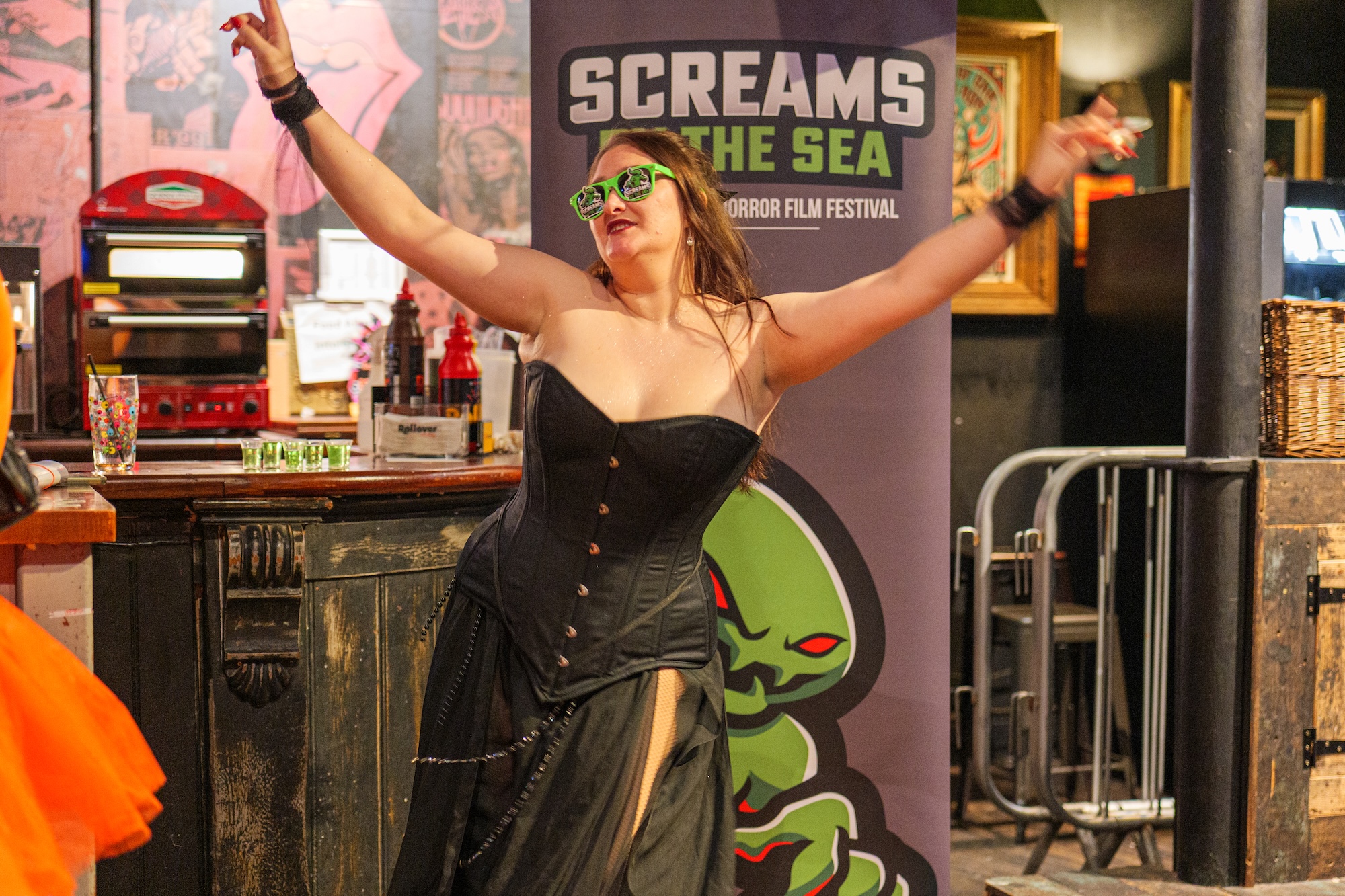 A person in a black corset dress and green sunglasses raises her arms at a horror film festival backdrop, with a poster reading 'SCREAMS ON THE SEA' behind her.