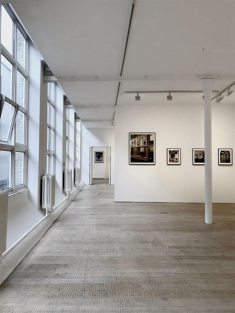 Interior of a gallery. A row of four artworks hang in frames on a wall to the right.