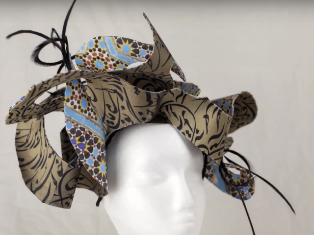 A woman's hat, made up of curves of gold and black fabric, sits on a mannequin's head
