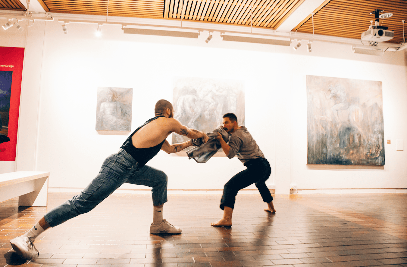 Two dancers performing a routine in a gallery in front of a row of artworks.