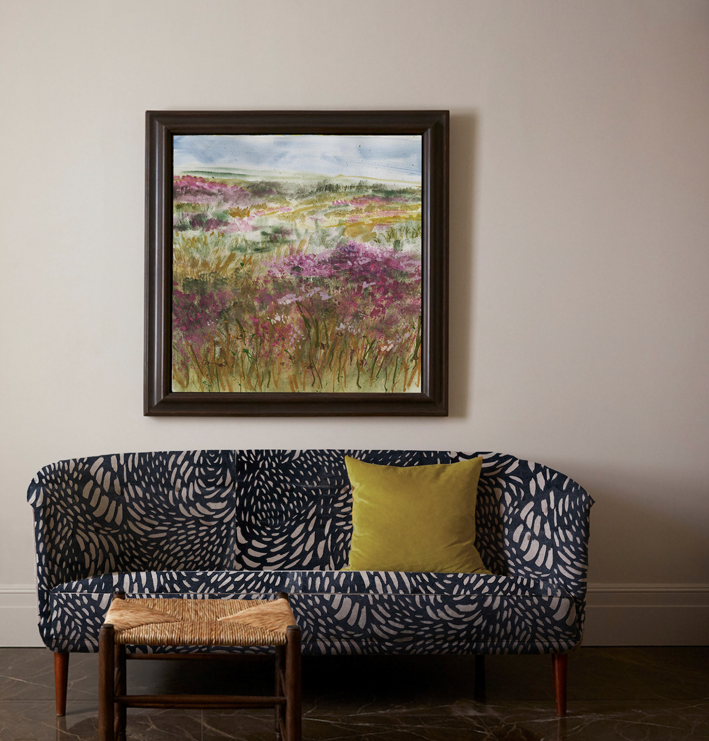 A sofa with feather-patterned upholstery. A wicker footstool is in front of it. A framed painting of wild flowers growing in a field hangs on the wall behind the sofa.