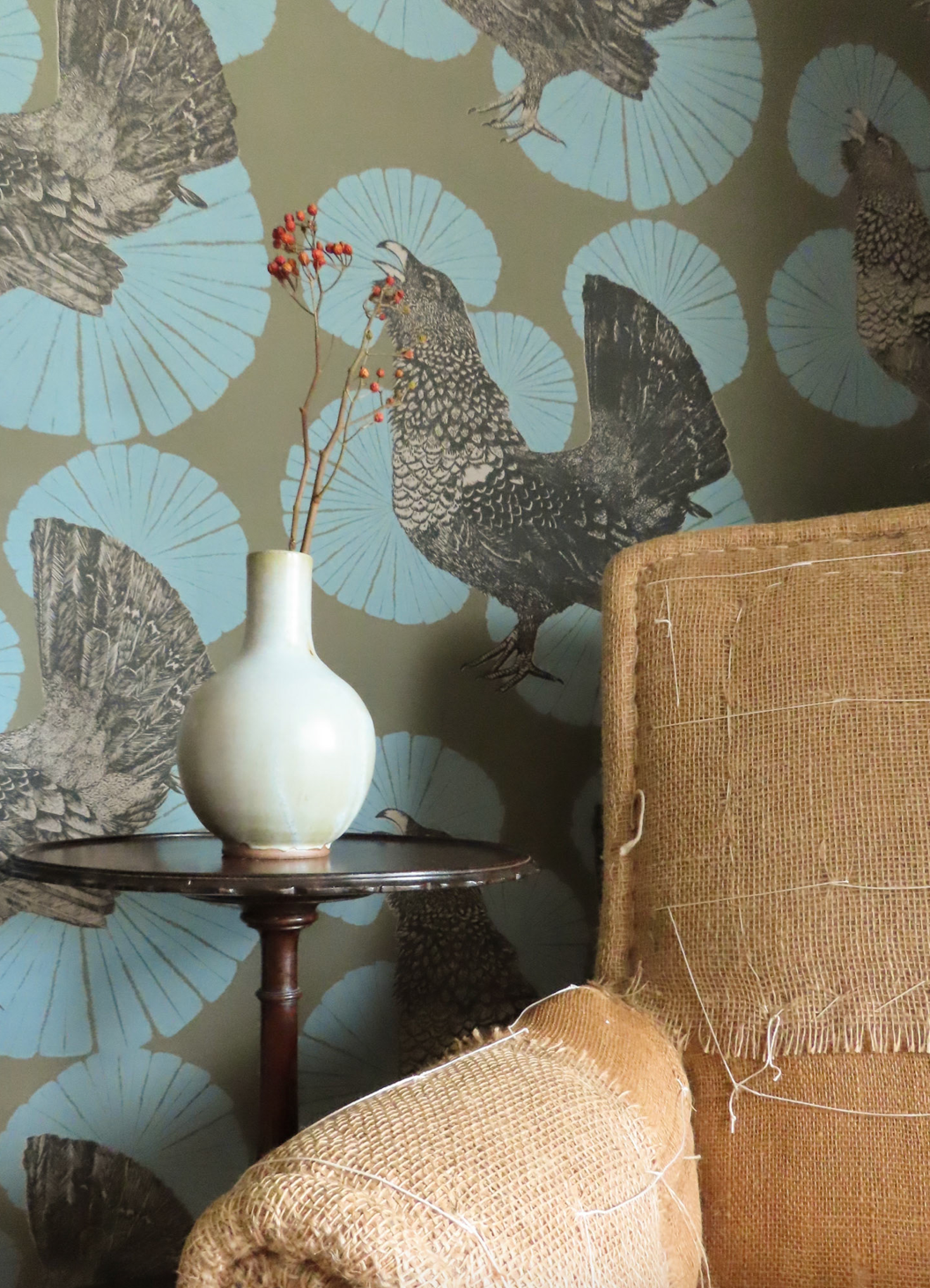 An armchair and a tea table with a small vase standing on it. The wallpaper is patterned with a repeated image of a chicken.