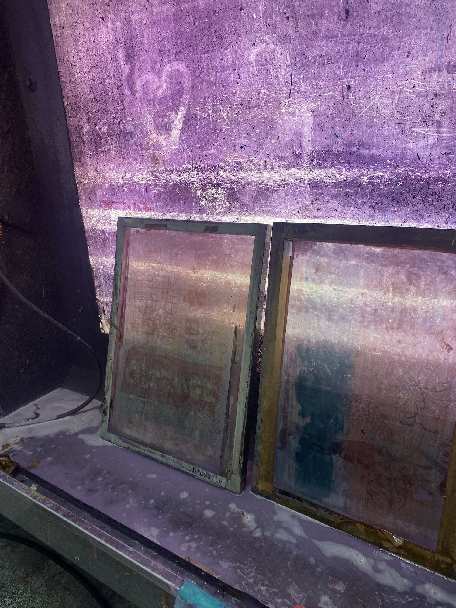 Two stained glass panes leaning against a purple wall, with a dusty ledge and dark curtain beside them.