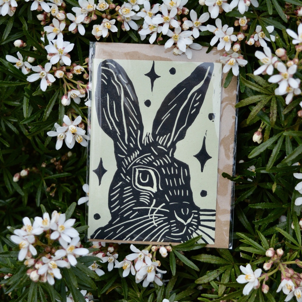 Card with a black screen print of a rabbit with sparkles on a green background, on top of a bed of white flowers and green foliage.