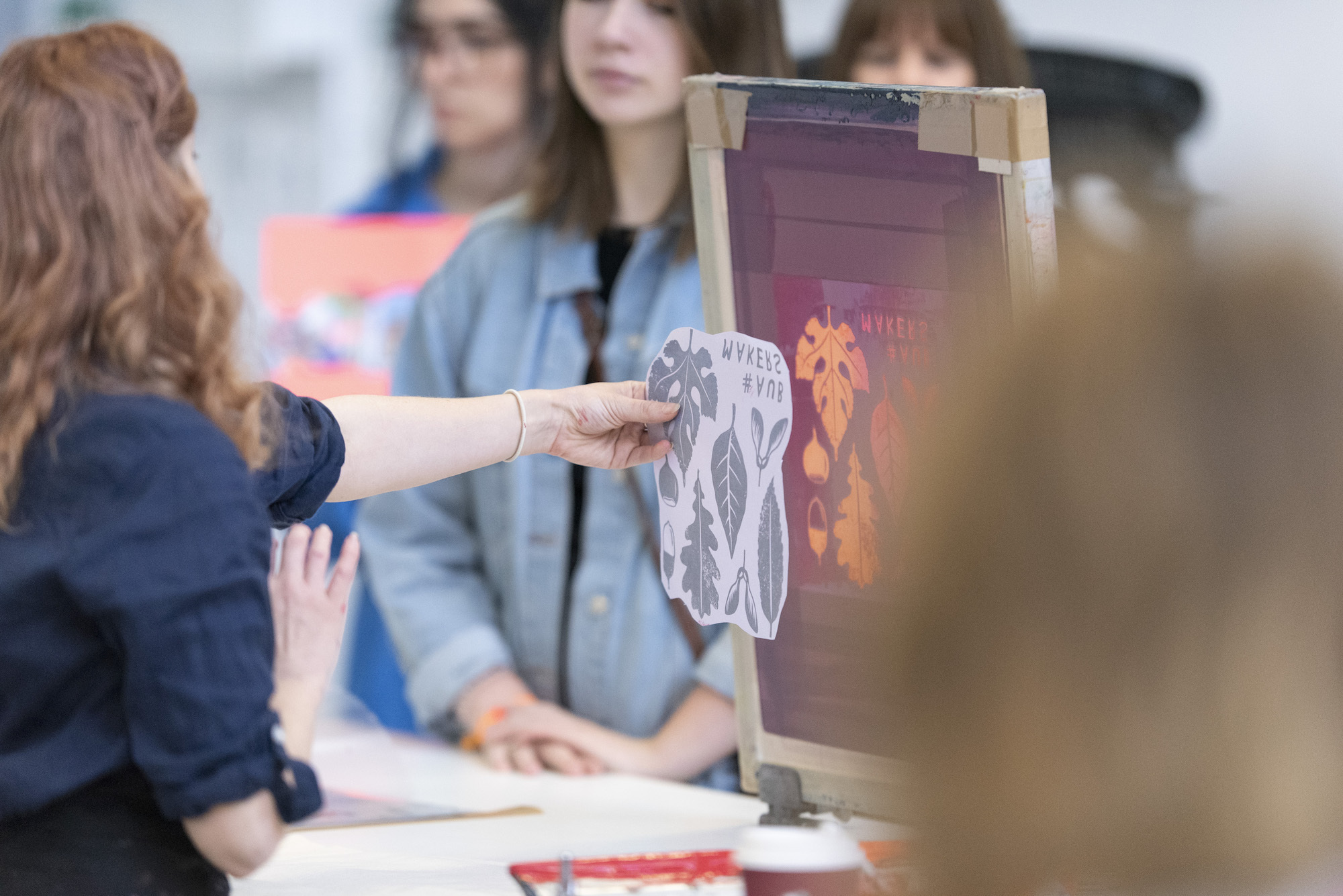 A person holds a piece of paper up to an identical screen printed image, demonstrating to students screen printing technique.