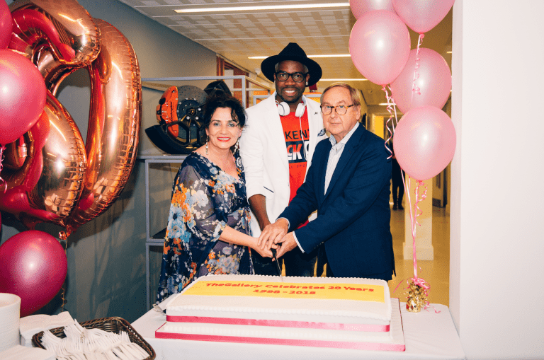 Three people hold a knife preparing to cut into a cake, surrounded by balloons.
