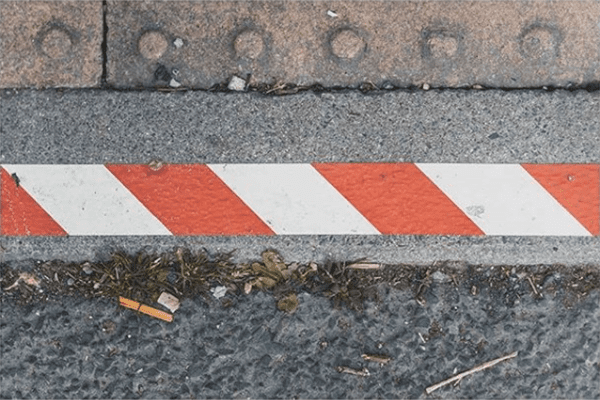 The edge of a pavement and road with a piece of red and white striped tape going across.