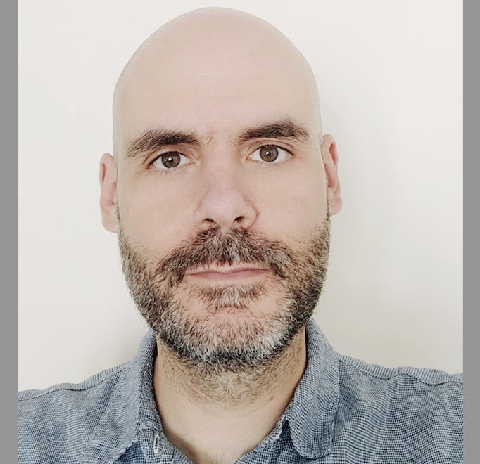 Bald man with brown beard wearing grey checked shirt against pale background, looking directly at camera.