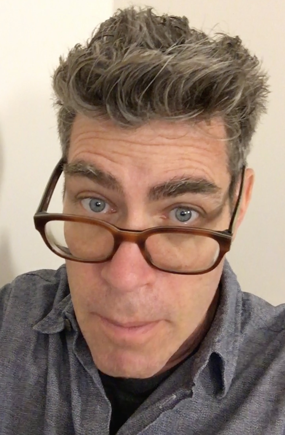 Man with brown-framed glasses peering over them, grey wavy hair, blue eyes, wearing grey shirt against cream background.