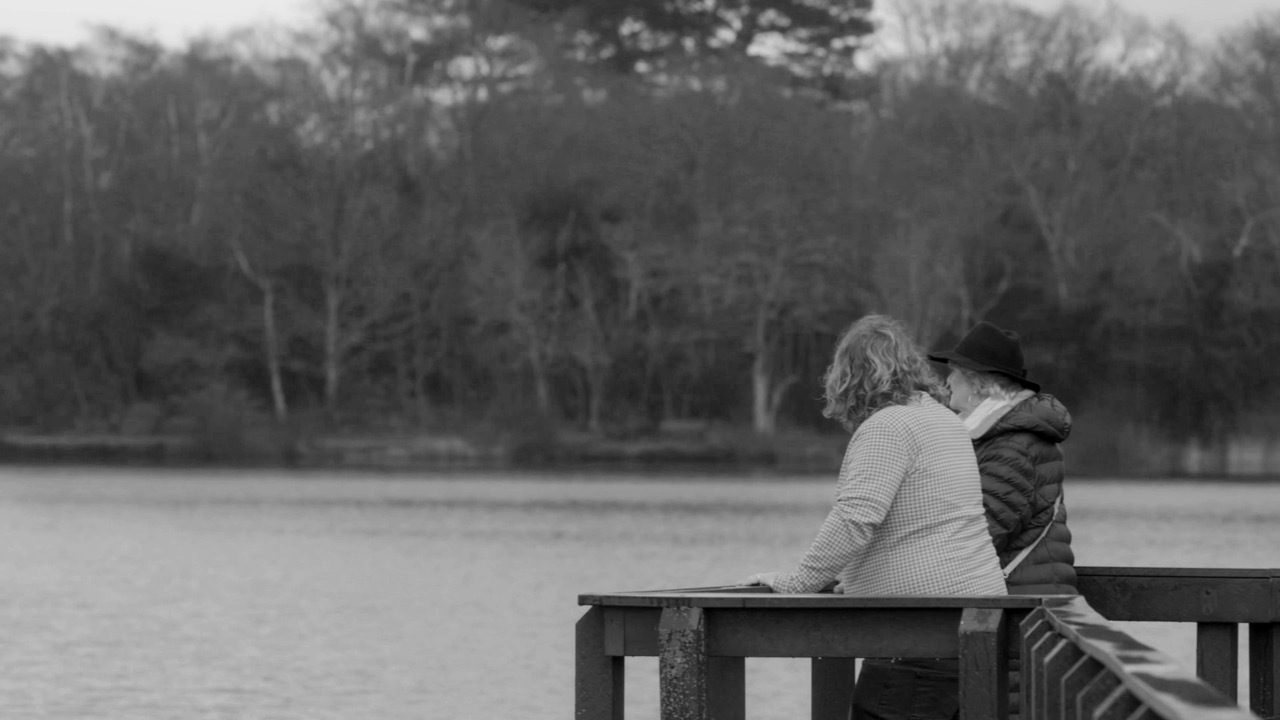 Black and white shot of two people stood by a lake surrounded by trees.