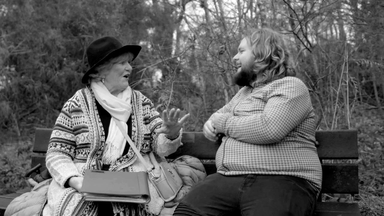 Black and white shot of Jacob Parvin sat on a bench with his grandmother, in conversation, trees in the background.