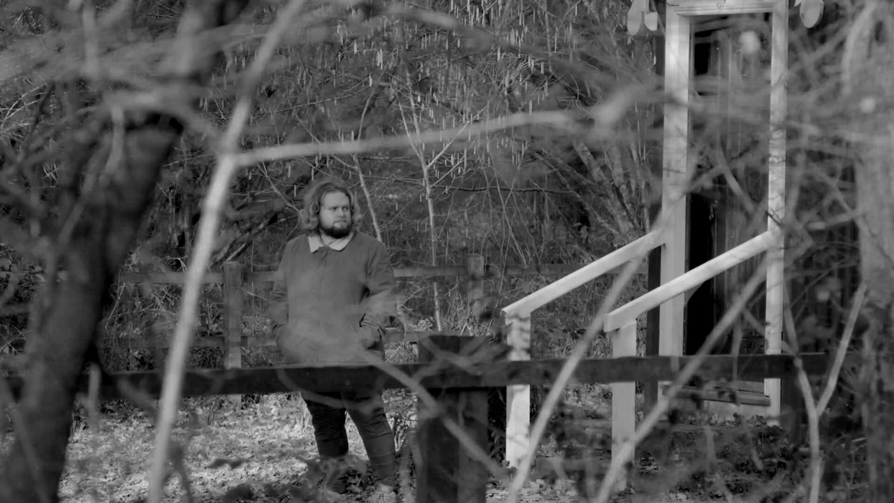 Black and white shot of Jacob Parvin walking through the woods looking at the entrance of a white frame building, blurred branches and trees.