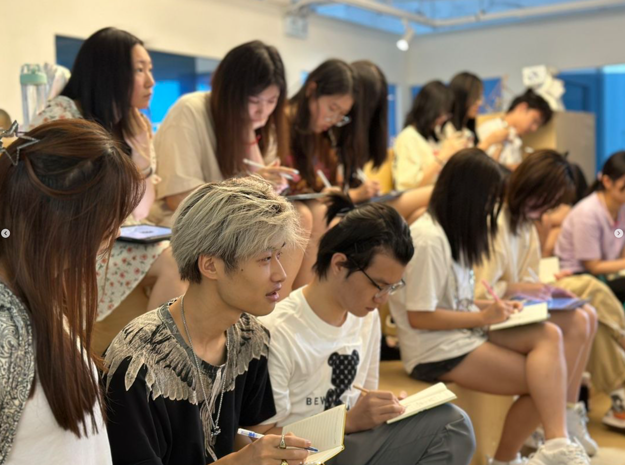 教室中的年轻学生，有人坐着、有人站着，正在纸上书写；前景人物留着银灰色头发，身穿流苏上衣。